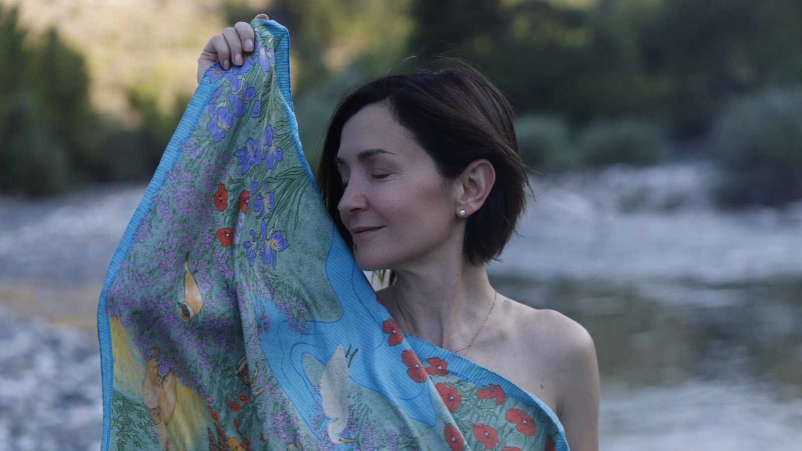 A woman with closed eyes gently holds a colorful silk scarf to her face by a riverside, symbolizing softness, self-care, and respect for the skin’s natural beauty.
